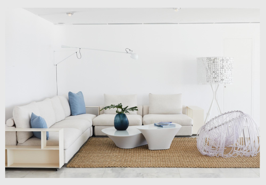 Designhotels, Myconian Kyma, Mykonos, Greece by Studio Linda Ehrl living room in with a white sofa, 2 blue pillows, a brown rug and a floor lamp in white.