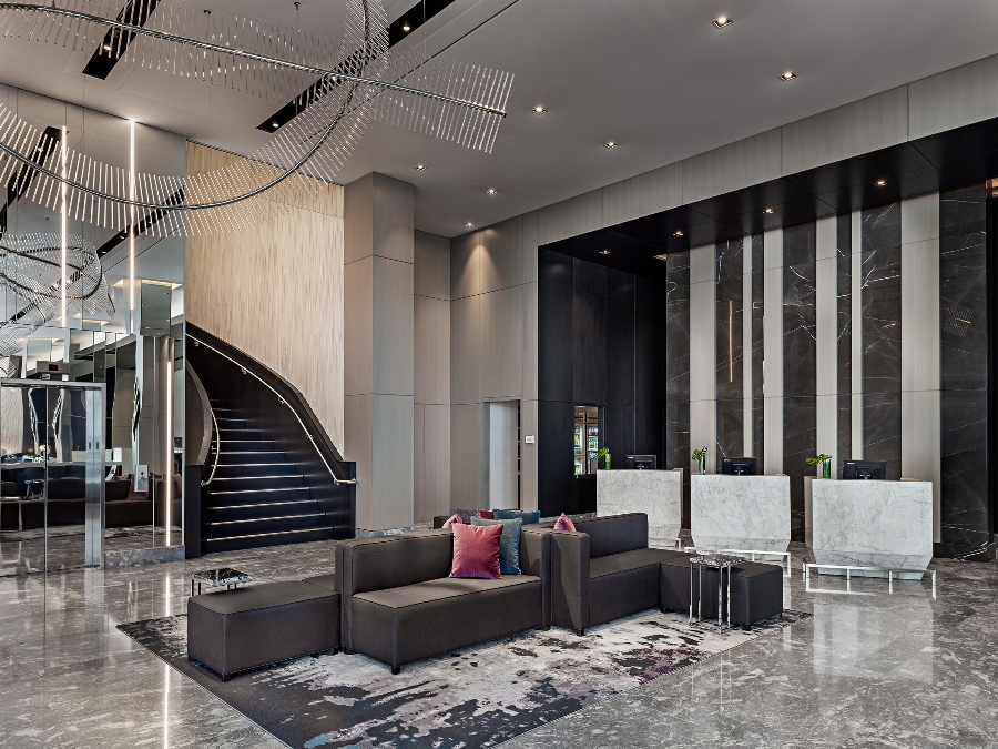 Hotel Decoration With II BY IV DESIGN. The lobby of the Toronto Marriott Markham has grey sofas in the center with pink and blue pillows and a grey and black rug.
